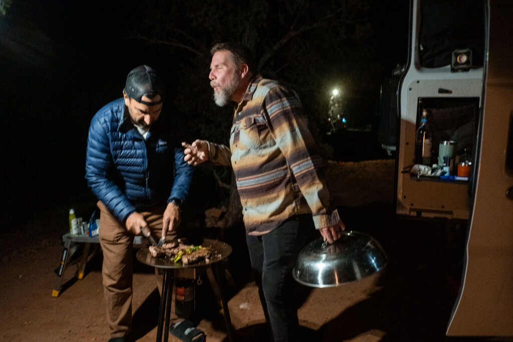 Two men are cooking outdoors at night, one slicing meat on a table while the other holds a large bowl. A vehicle is nearby.